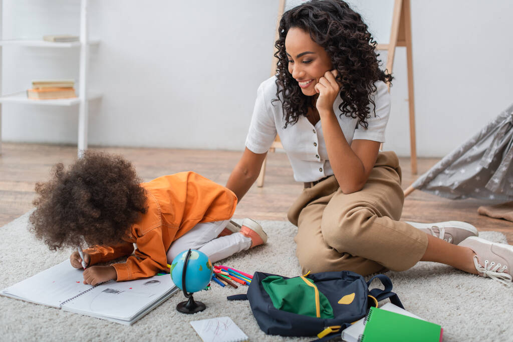 Усміхаючись африканською американською жінкою сидячи біля маленької дочки малюючи на записнику вдома.  - Фото, зображення