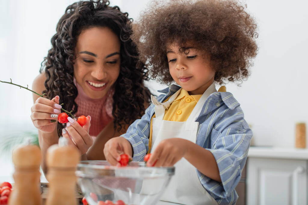Toddler african american kid holding cherry tomatoes near smiling mom in kitchen  - 写真・画像