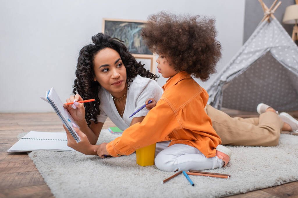 Afričanky americká žena drží notebook v blízkosti dcery a barevné tužky na koberec doma  - Fotografie, Obrázek