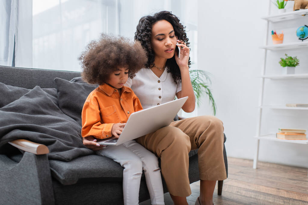 Afroamerykanka rozmawia na smartfonie w pobliżu córki za pomocą laptopa na kanapie  - Zdjęcie, obraz