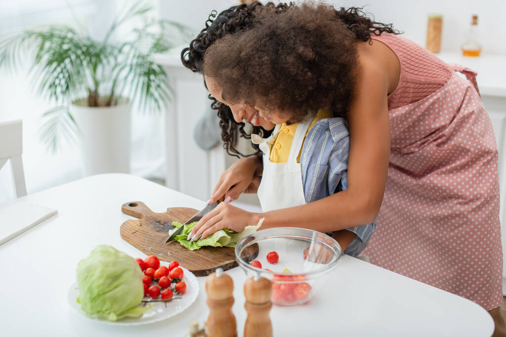 Jovem mãe afro-americana cortando repolho perto de criança e tomates cereja na cozinha  - Foto, Imagem
