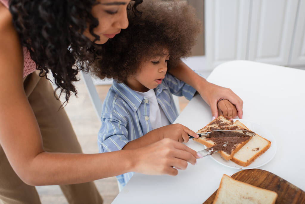 afro-américaine femme et enfant propagation pâte de chocolat sur le pain dans la cuisine  - Photo, image