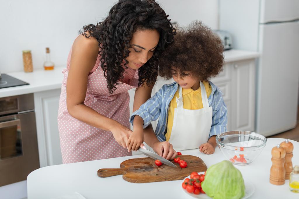 Молодая африканская американка режет помидоры черри рядом с дочерью и капустой на кухне  - Фото, изображение