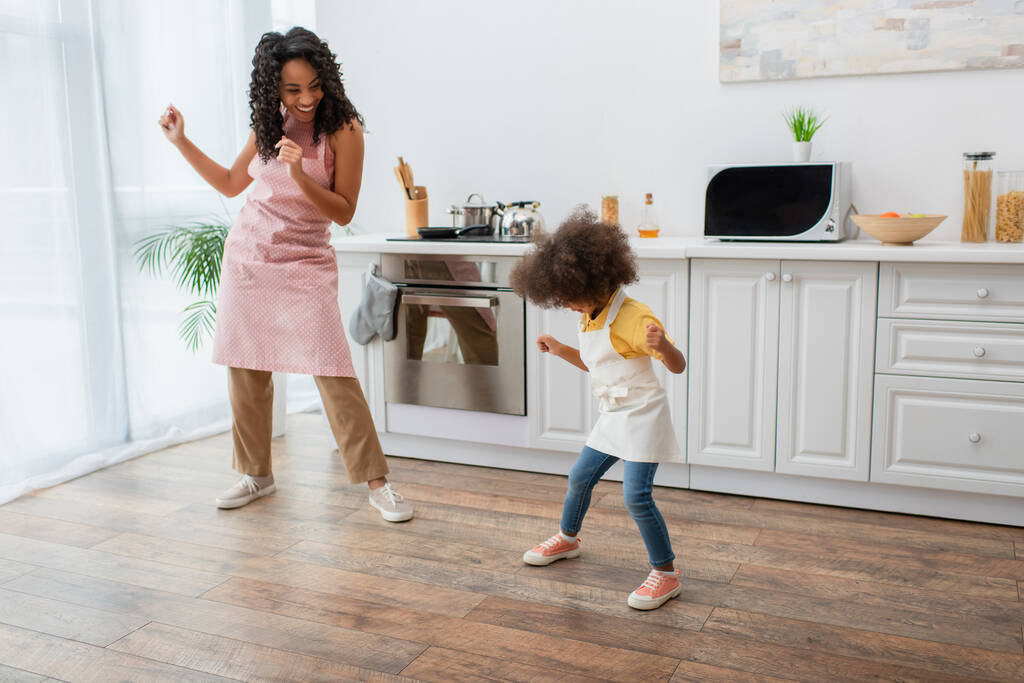 Felice donna afroamericana e bambino in grembiuli che ballano in cucina  - Foto, immagini