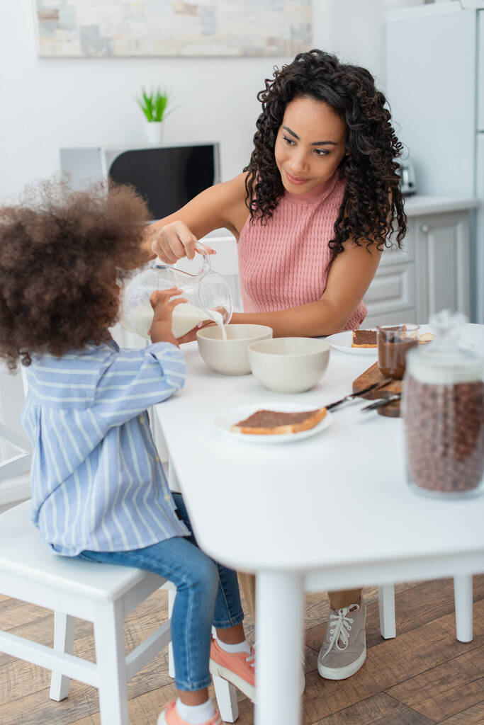 Jeune mère afro-américaine versant du lait près de sa fille et petit déjeuner dans la cuisine  - Photo, image