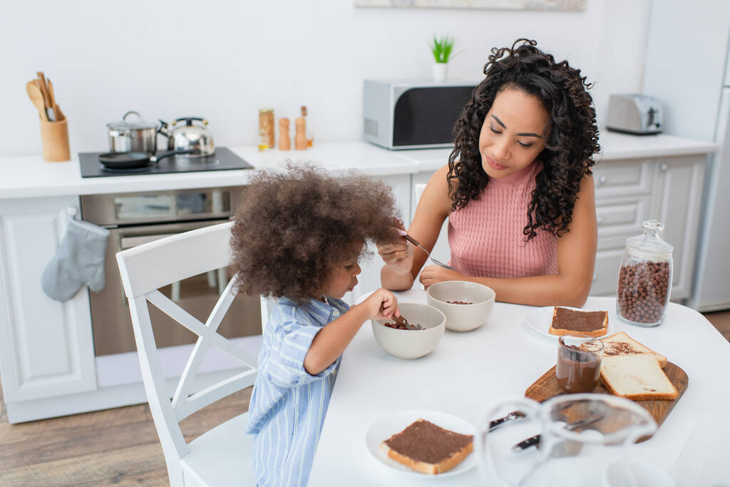 Mujer afroamericana joven sosteniendo cuchara cerca de niño con cereal y pan en la cocina  - Foto, imagen
