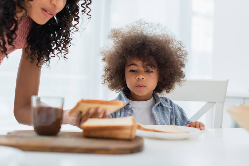 アフリカ系アメリカ人の子供近くに座っているお母さんとパンとぼやけたチョコレートペーストでキッチン  - 写真・画像