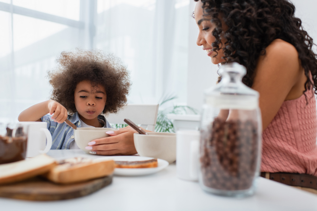 Mère afro-américaine tenant bol près de sa fille et petit déjeuner flou à la maison  - Photo, image