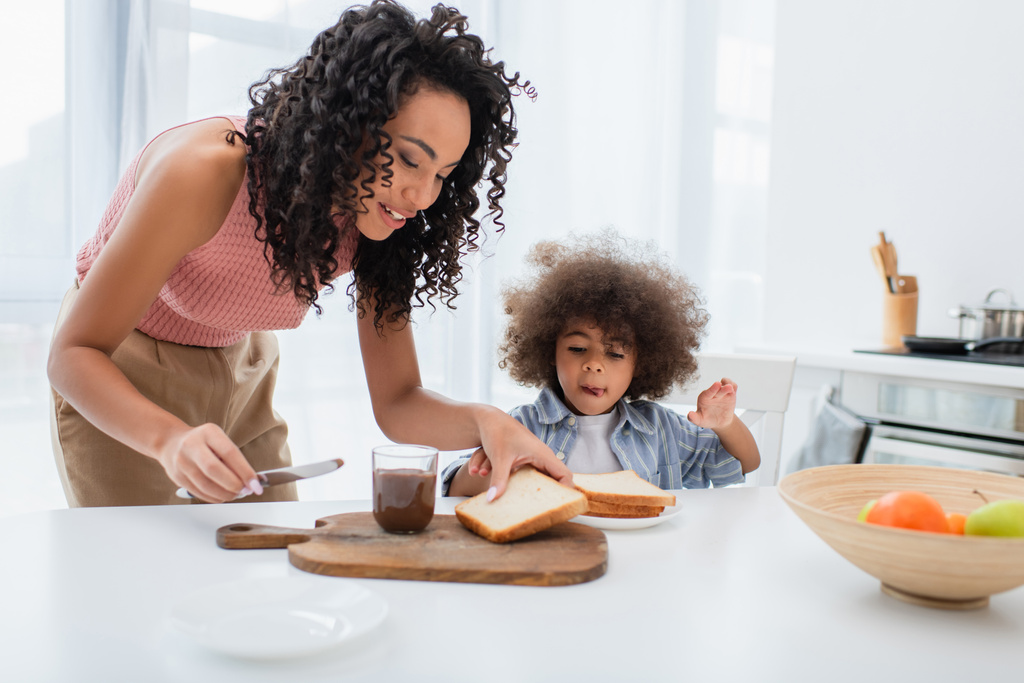 笑顔アフリカ系アメリカ人女性保持ナイフとパン近く娘と果物でキッチン  - 写真・画像