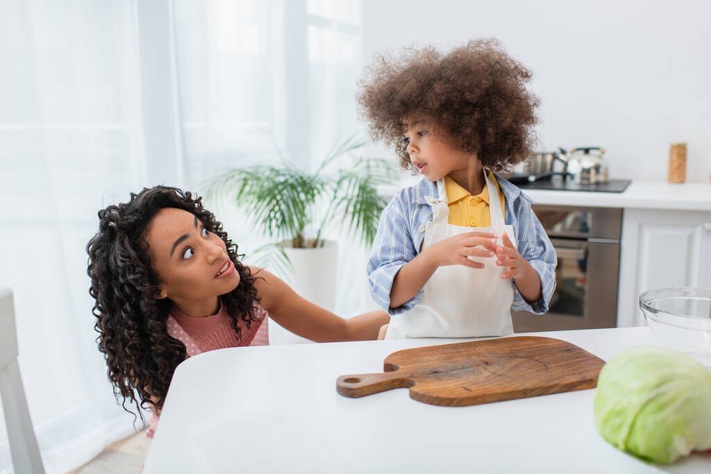 Mujer afroamericana asustada mirando a su hija pequeña cerca de repollo y tabla de cortar en la cocina  - Foto, imagen