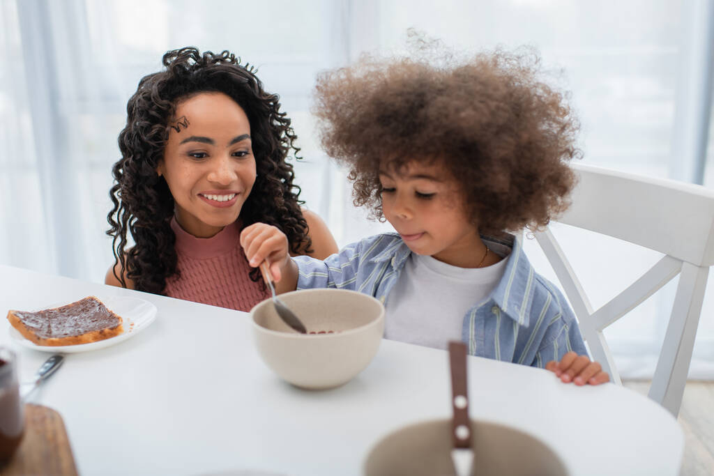 Feliz mujer afroamericana mirando a su hija con cereal en la cocina  - Foto, imagen