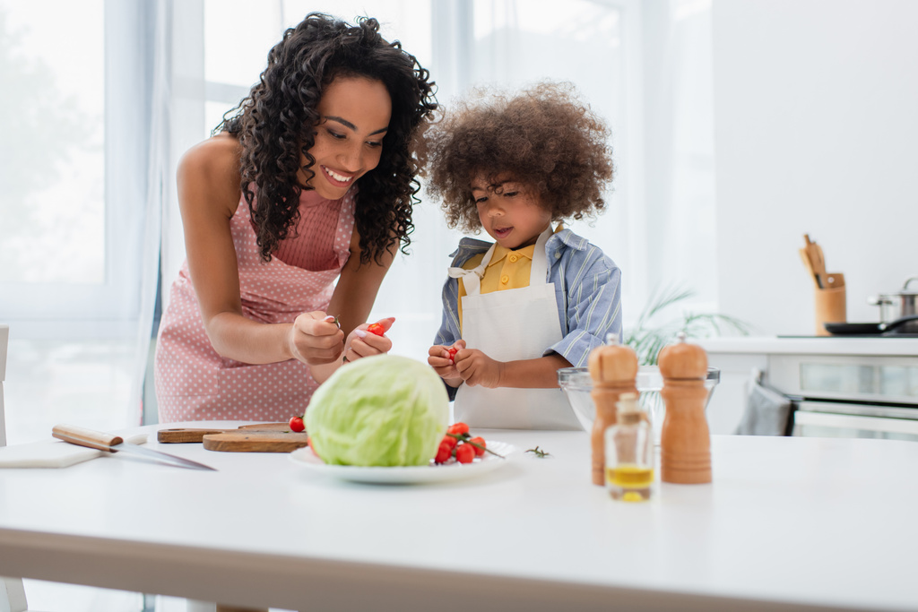 Önlüklü Afro-Amerikan bir aile, ellerinde kiraz domatesleri var. Mutfaktaki kesim tahtasının yanında.  - Fotoğraf, Görsel