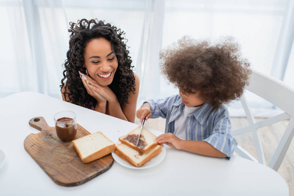 Χαμογελώντας αφρικάνικη αμερικανική μητέρα κοιτάζοντας το παιδί εξαπλώνεται πάστα σοκολάτας στο ψωμί στην κουζίνα  - Φωτογραφία, εικόνα