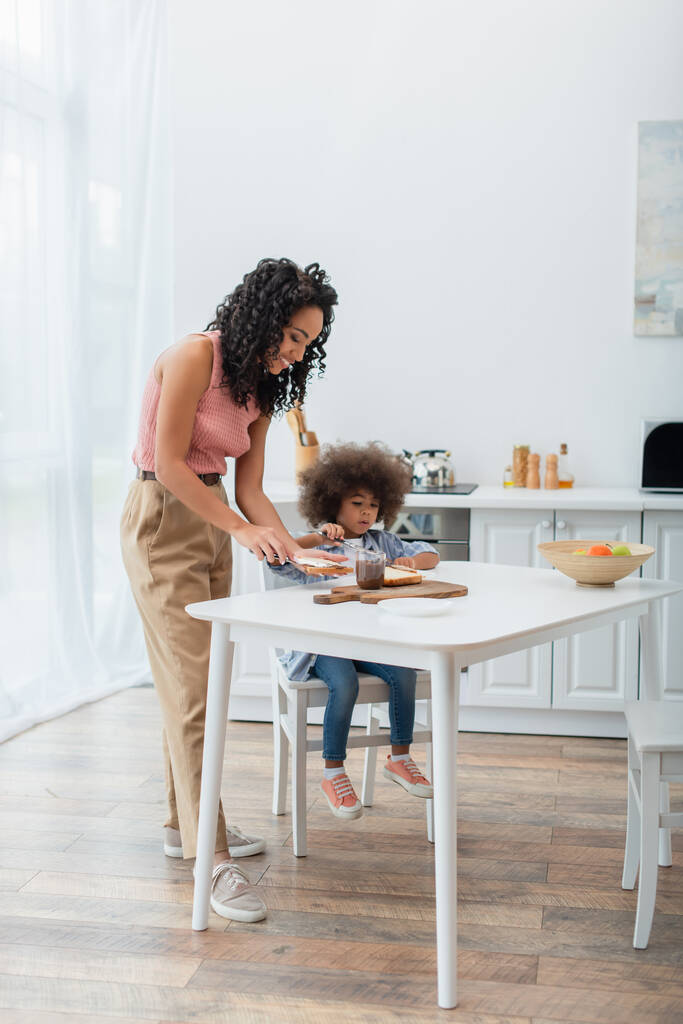 Jeune femme afro-américaine étalant de la pâte de chocolat sur du pain près d'un enfant et des fruits dans la cuisine  - Photo, image