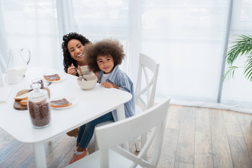Mosolygó afro-amerikai gyerek néz kamera közelében anya és reggeli a konyhában  - Fotó, kép
