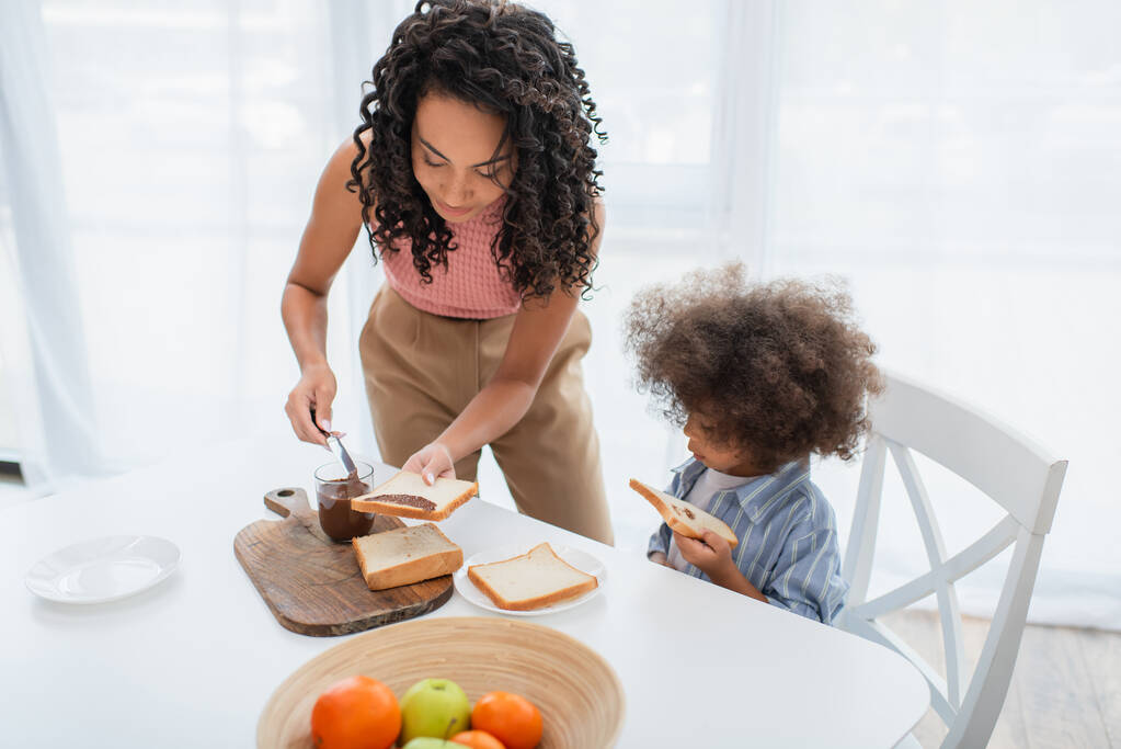 Genç Afrikalı Amerikalı anne çocuğun yanına çikolata ezmesi sürüyor ve mutfakta meyveler var.  - Fotoğraf, Görsel