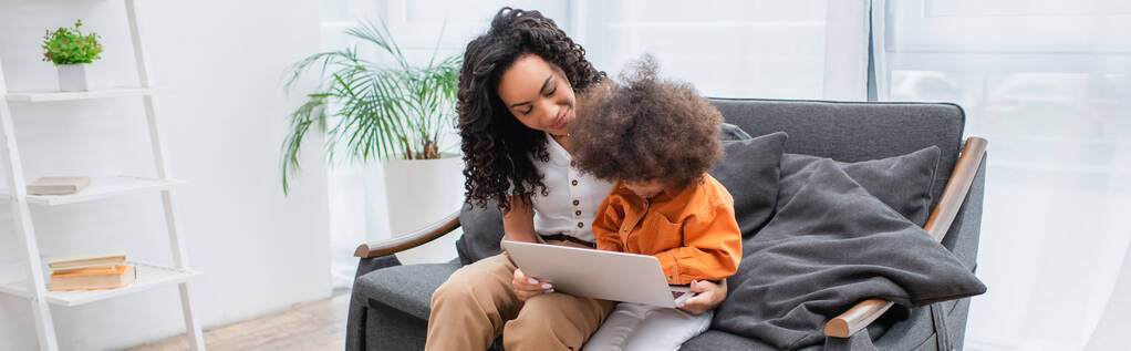 Sonriente madre afroamericana sosteniendo portátil cerca de niño en el sofá en casa, pancarta  - Foto, imagen