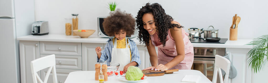 Bambino afroamericano in grembiule in piedi vicino alla mamma e verdure fresche in cucina, banner  - Foto, immagini