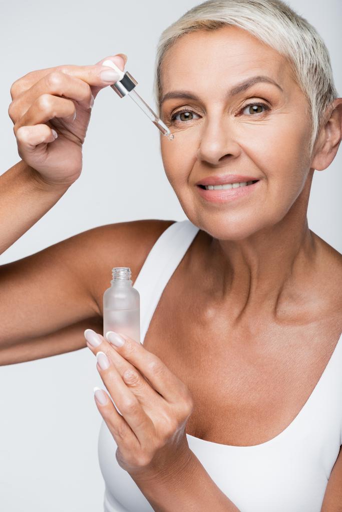 gelukkig senior vrouw aanbrengen serum met pipet geïsoleerd op grijs - Foto, afbeelding