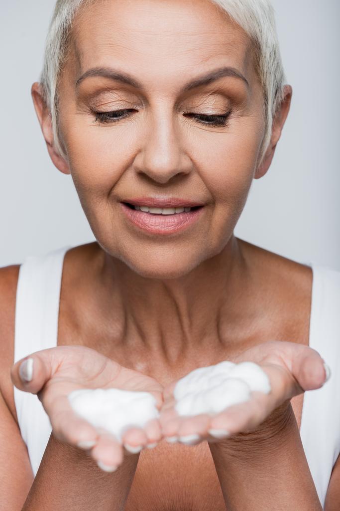 felice donna anziana guardando schiuma detergente sulle mani isolate su grigio  - Foto, immagini
