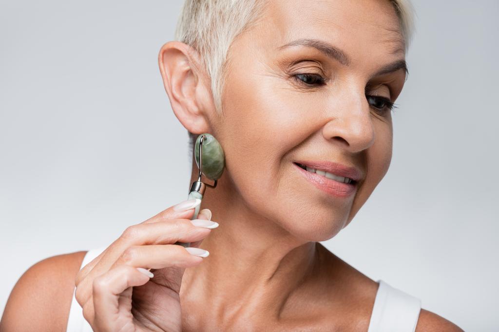 cara de masaje mujer senior positiva con rodillo de jade aislado en gris - Foto, Imagen