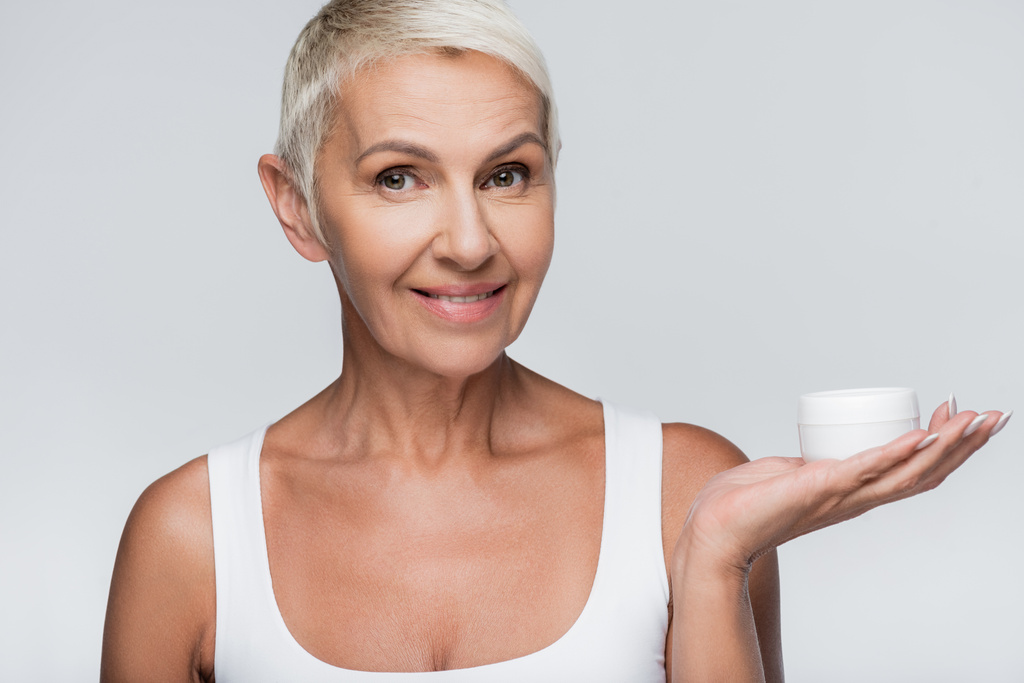 glückliche Seniorin zeigt Container mit Kosmetikcreme isoliert auf grau - Foto, Bild