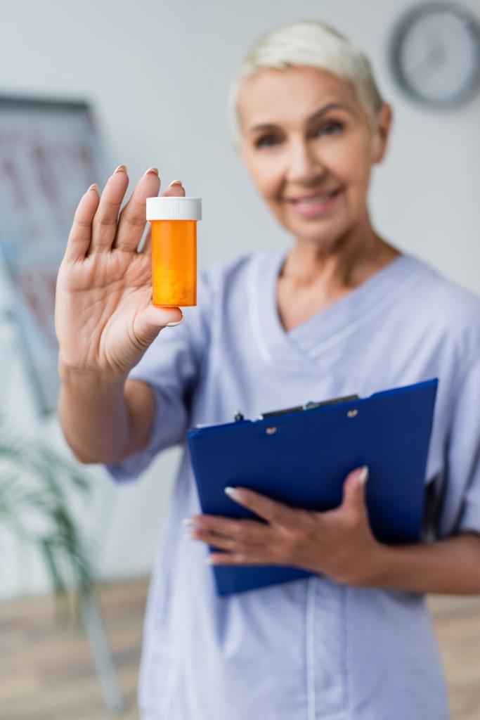 enfermera sonriente y borrosa sosteniendo botella con pastillas y portapapeles  - Foto, imagen