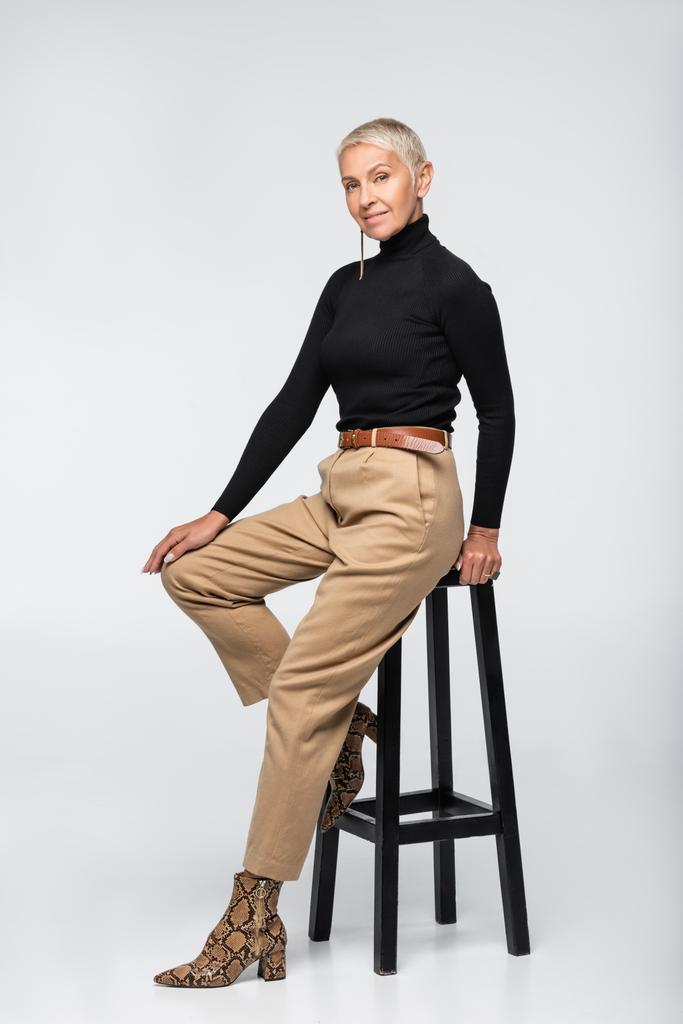 full length of pleased senior woman in beige pants and turtleneck posing near chair on grey - Photo, Image