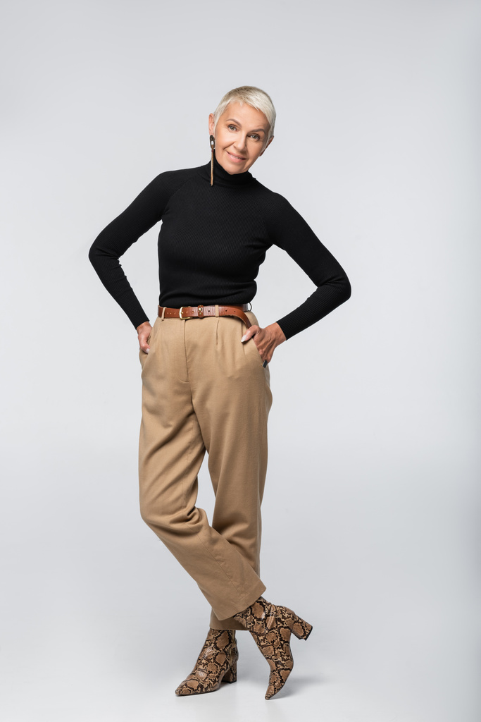 longueur totale de femme âgée souriante en pantalon beige, col roulé noir et bottes à imprimé animal posant avec les mains dans les poches sur gris - Photo, image