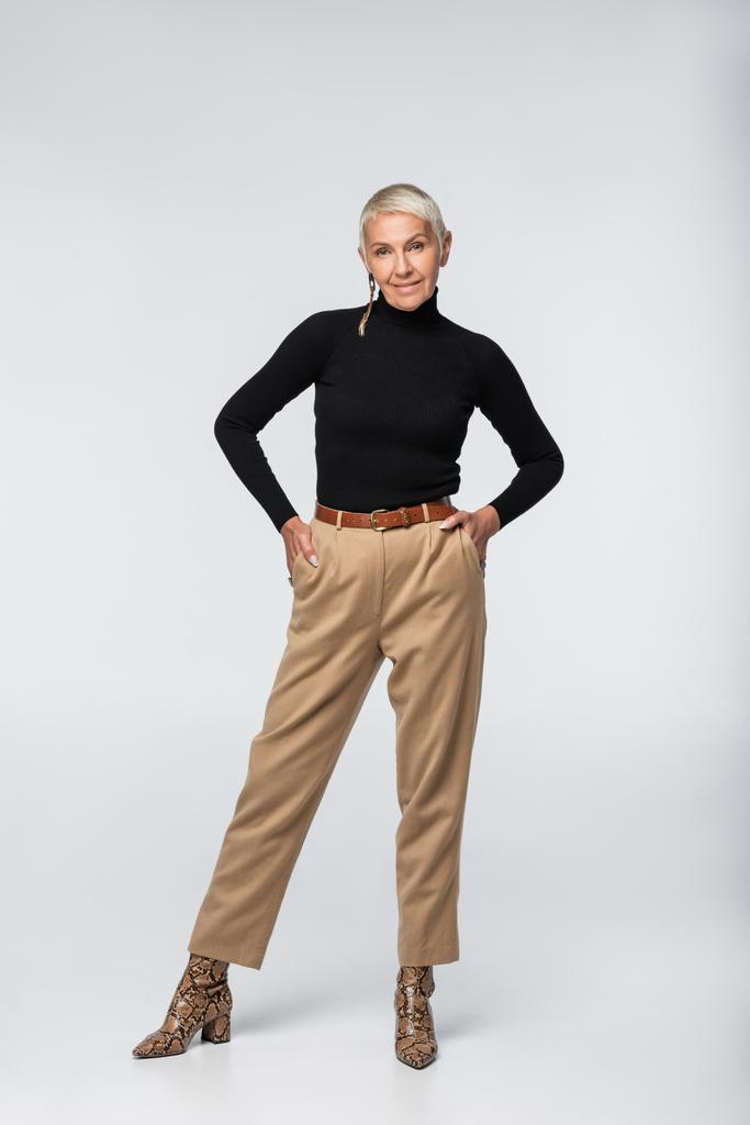 pełna długość zadowolonej i modnej starszej kobiety w spodniach, czarnym golfie i zwierzęcych butach z nadrukiem, pozujących z rękami w kieszeniach na szarości - Zdjęcie, obraz