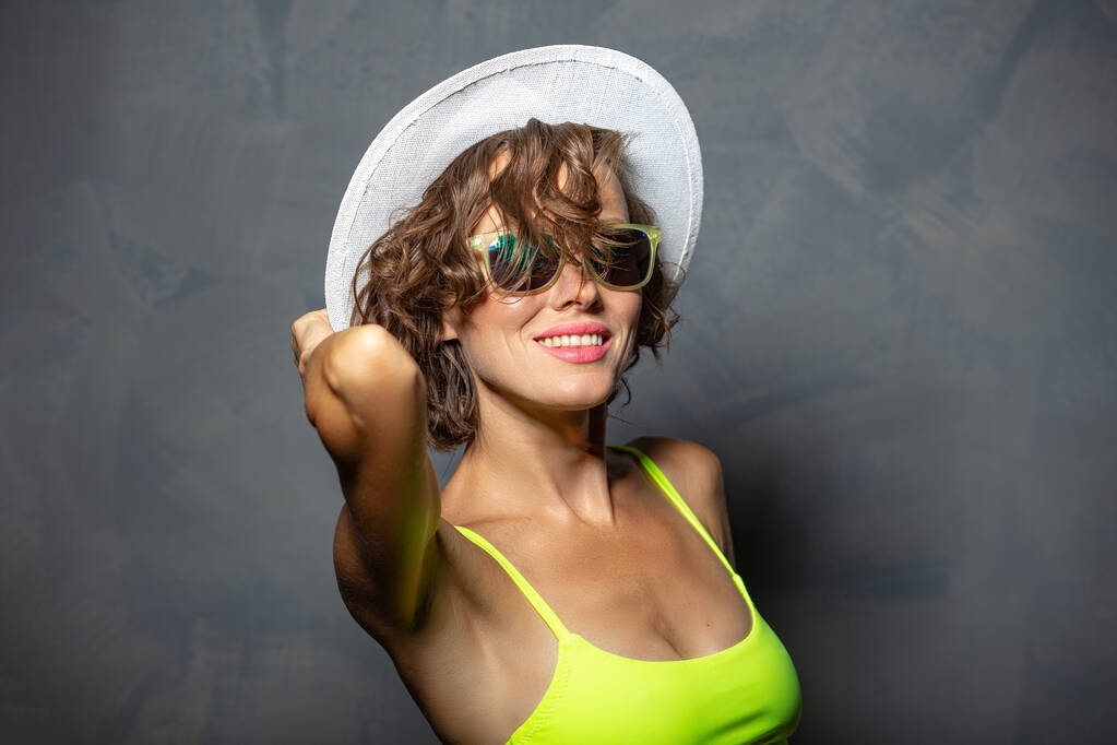 Красивая брюнетка в цветном купальнике и летающих волосах, копия пространства в шляпе и солнцезащитных очках - Фото, изображение