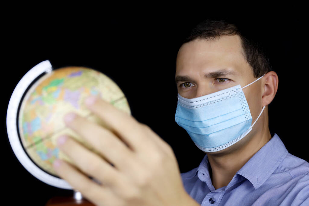 Turismo durante a pandemia de coronavírus, homem de máscara protetora olhando para o globo. Escolhendo passeio de viagem, retrato masculino no fundo preto - Foto, Imagem