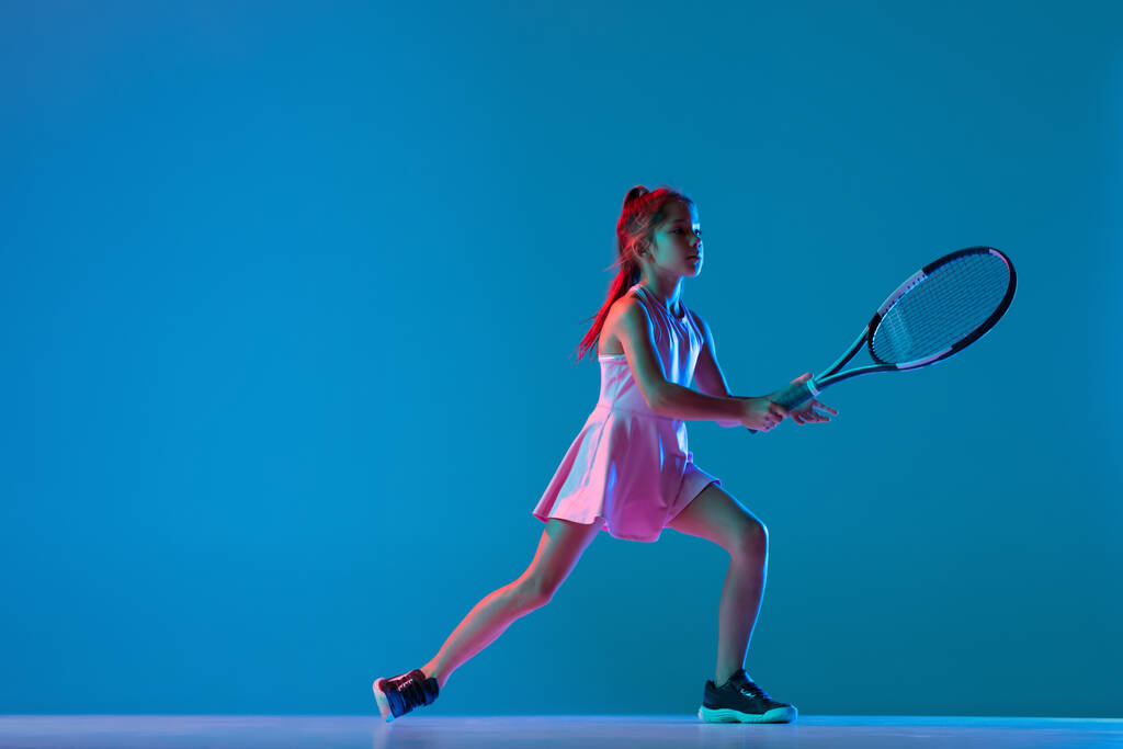 Πορτρέτο του μικρού κοριτσιού, αρχάριος παίκτης του τένις παίζει τένις απομονωμένο σε μπλε φόντο στούντιο σε νέον φως. Αθλητισμός, μελέτη, παιδική ηλικία - Φωτογραφία, εικόνα