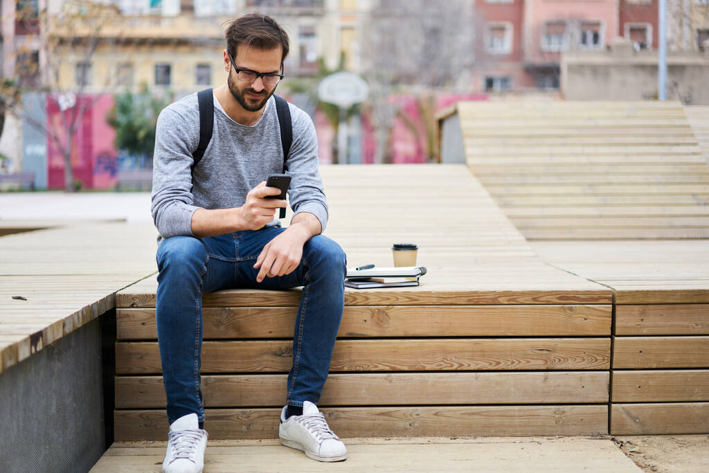 Brodaty mężczyzna z plecakiem i na wynos gorąca kawa siedzi na ławce i sprawdza e-mail na smartfonie podczas spędzania czasu na ulicy - Zdjęcie, obraz