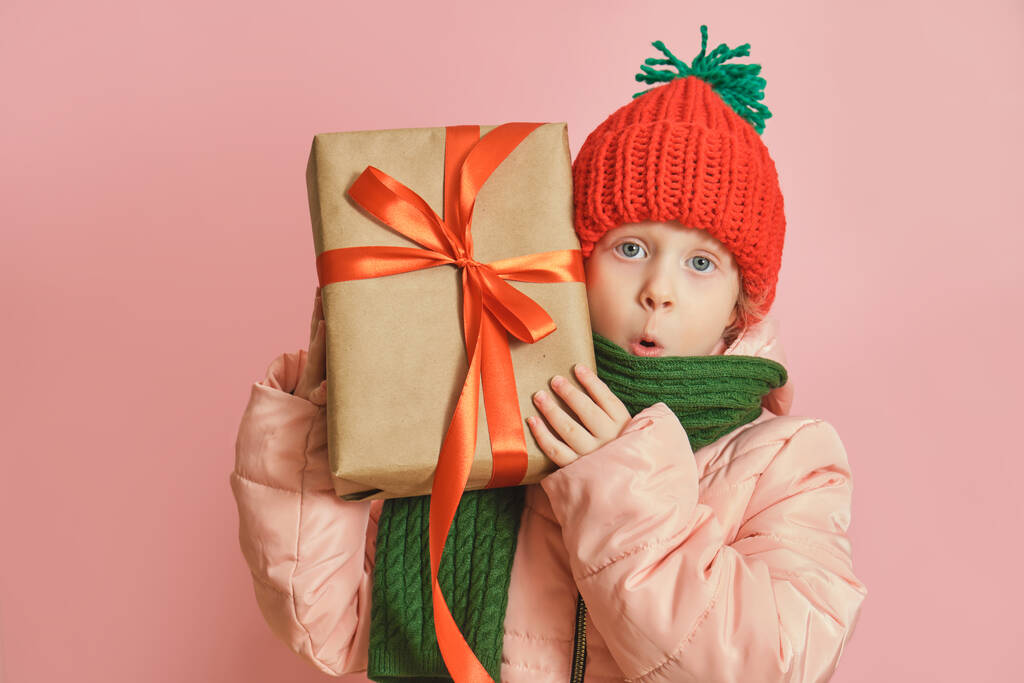 Boldog aranyos kaukázusi kis mosolygós kislány karácsonyi ajándékdobozzal piros szalaggal. lány kötött sál és kalap rózsaszín háttér,, karácsonyi hangulat koncepció másolás tér - Fotó, kép