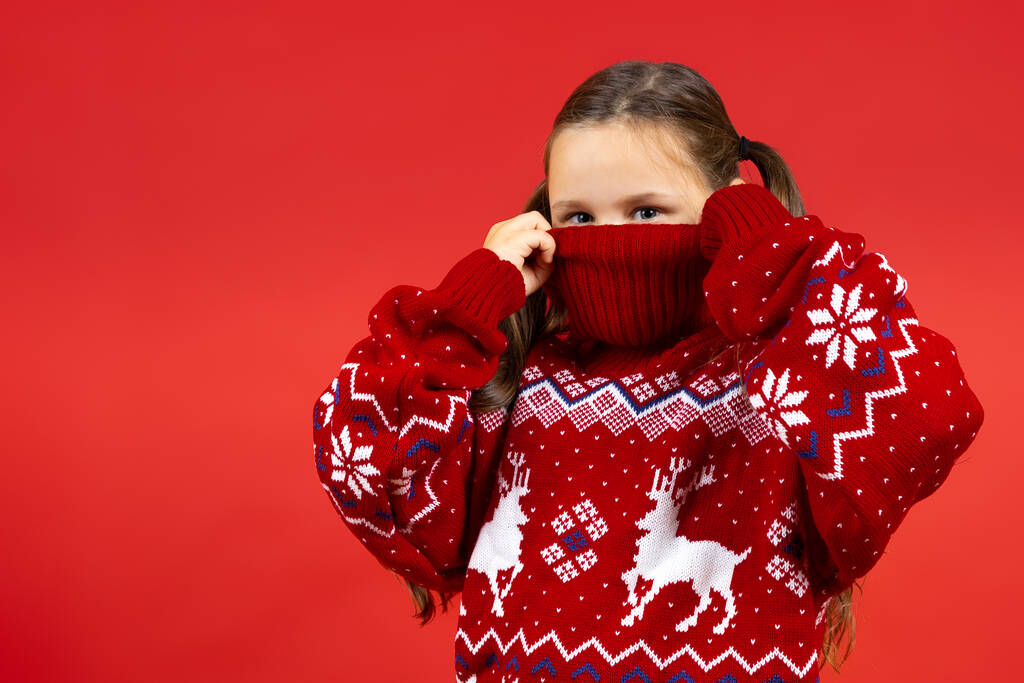 portrait rapproché de fille ludique cachant demi-visage en pull de Noël rouge avec des rennes, isolé sur fond rouge - Photo, image