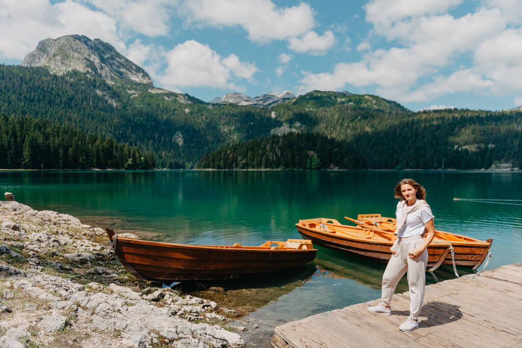 Kız, Karadağ Durmitor Ulusal Parkı 'ndaki balıkçı tekneleri, ormanlar ve dağlarla Kara Göl' e bakıyor. Bir turist sihirli bir göl manzarası, kozalaklı bir orman ve büyülü bir manzaranın tadını çıkarır. - Fotoğraf, Görsel