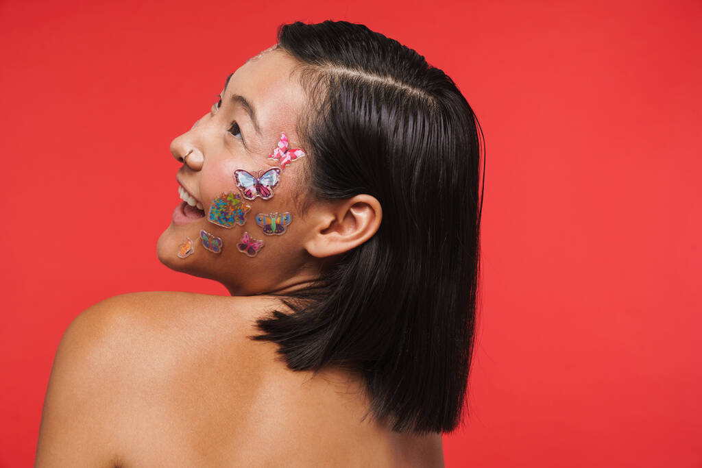 Retrato de belleza de una joven morena feliz sonriente asiática con maquillaje facial decoración posando sobre fondo de pared roja vista posterior - Foto, Imagen