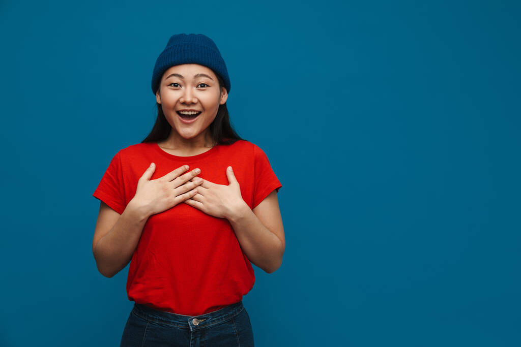Muotokuva onnellinen innoissaan rento nuori aasialainen nainen yllään hattu seisoo sininen seinä tausta kädestä rinnassa - Valokuva, kuva