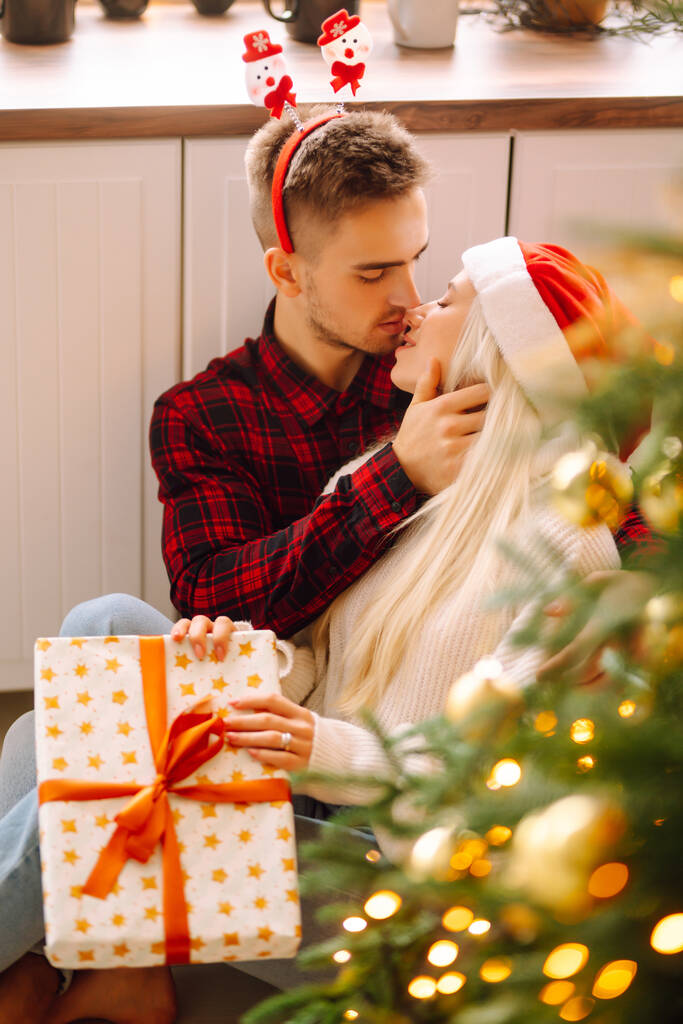 Νεαρό ζευγάρι με χριστουγεννιάτικα δώρα στο σπίτι κοντά στο χριστουγεννιάτικο δέντρο. Ρομαντική μέρα. Χειμερινές διακοπές. Πρωτοχρονιά. - Φωτογραφία, εικόνα