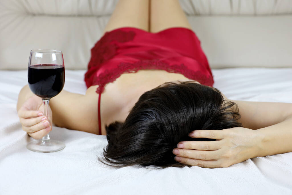 Alusvaatteinen nainen makaa punaviinin kanssa sängyllä. Juhlan, rentoutumisen, naisten alkoholismin käsite - Valokuva, kuva
