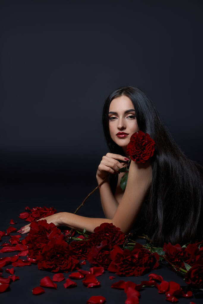 Όμορφη μελαχρινή γυναίκα με ένα μπουκέτο τριαντάφυλλα λουλούδια κάθεται στο πάτωμα σε ένα μαύρο κοστούμι. Μακριά μαλλιά, λεπτό σώμα - Φωτογραφία, εικόνα