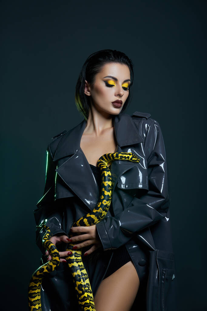 Beauty woman python yellow snake around her neck on latex shiny raincoat. Yellow snake on the shoulders of girl. Beauty yellow eye shadow makeup, dark burgundy lipstick - Photo, Image