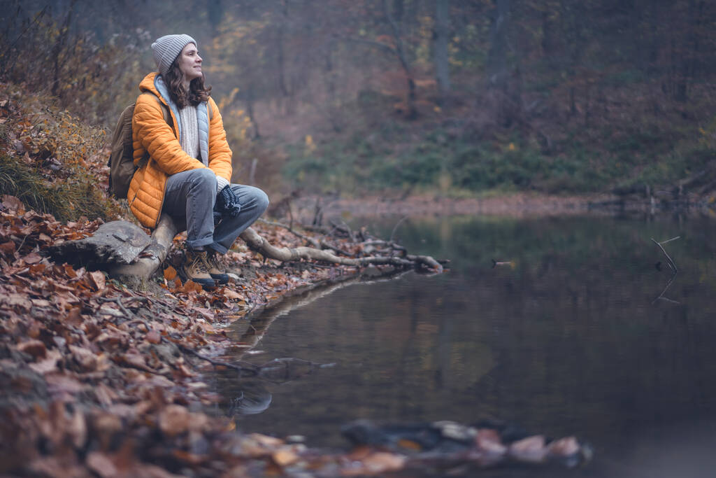 Mujer joven viajera con una chaqueta amarilla y sombrero sentado en un tronco junto al estanque en el bosque nublado de otoño - Foto, Imagen