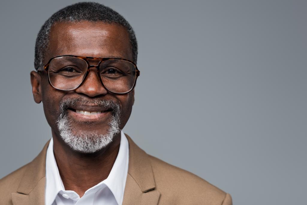 vousatý africký americký podnikatel v brýlích s úsměvem na kameru izolované na šedé - Fotografie, Obrázek