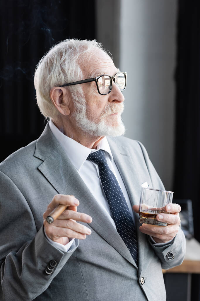 Älterer Geschäftsmann mit Zigarre und Glas Whiskey schaut im Büro weg - Foto, Bild