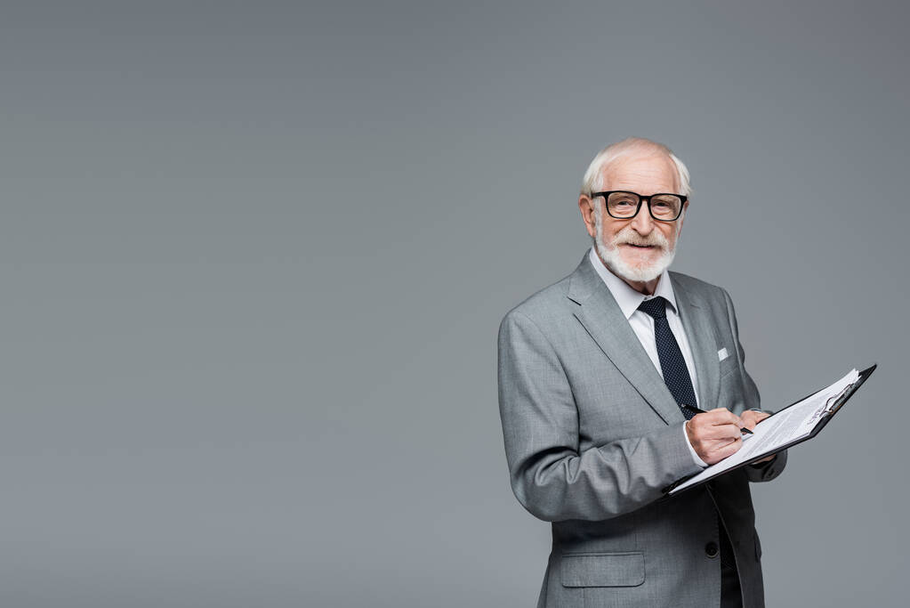 улыбающийся старший бизнесмен смотрит в камеру, подписывая контракт, изолированный от серых - Фото, изображение
