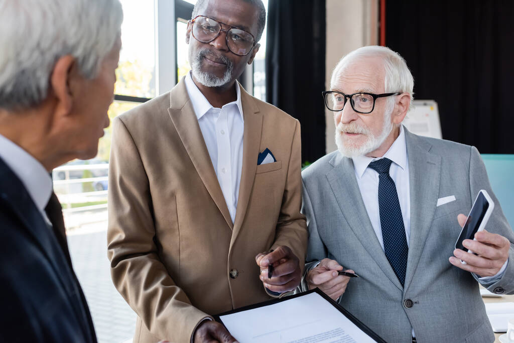 senior hommes d'affaires multiethniques montrant contrat et smartphone avec écran blanc à un collègue flou - Photo, image