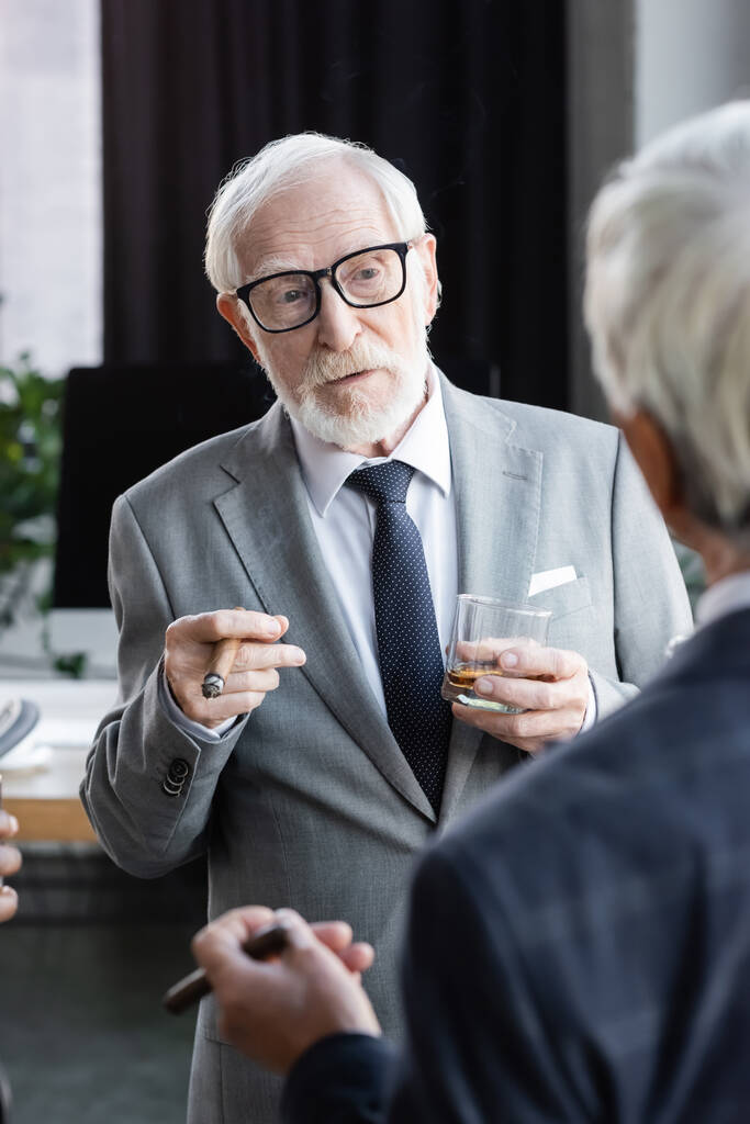 elderly businessman in eyeglasses holding cigar and glass of whiskey near colleague on blurred foreground - Photo, Image