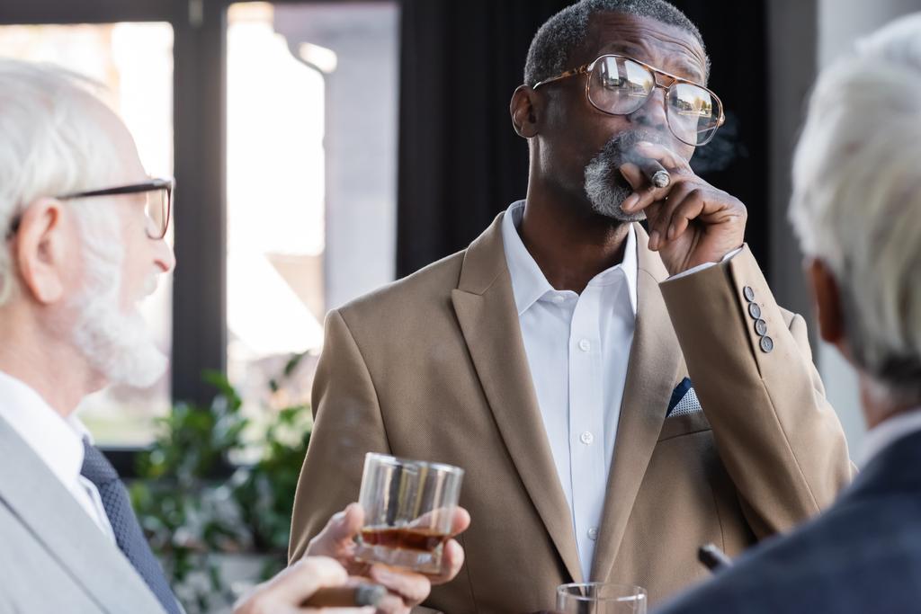 starszy afrykański amerykański biznesmen w okularach palący cygaro w pobliżu rozmazanych kolegów ze szklankami whisky - Zdjęcie, obraz